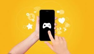 A hand holding and touching the screen of smartphone with game joystick on screen with award and achievement icons on yellow background. Gamification concept