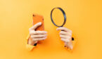 Hand holding magnifying glass and phone protruding from background